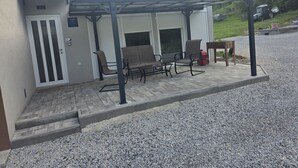 A covered outdoor seating area with a patio table and chairs, a door with a window, and a gravel driveway. - Vacation Home Rose (Kutjevo)