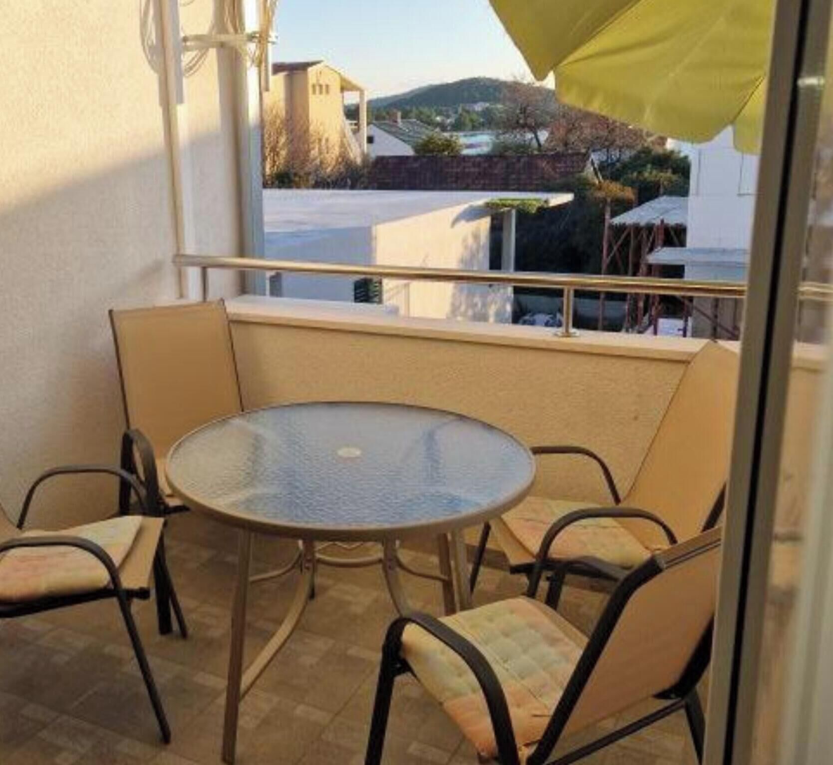 Ein Balkon mit rundem Glastisch, vier Stühlen, einem gelben Sonnenschirm und Blick auf Häuser und Hügel.