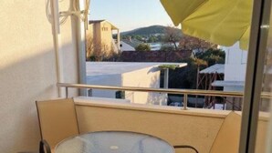 A balcony with a round glass table and four chairs, a yellow umbrella, and a view of buildings and hills. - Apartments Ordulj (Rogoznica)