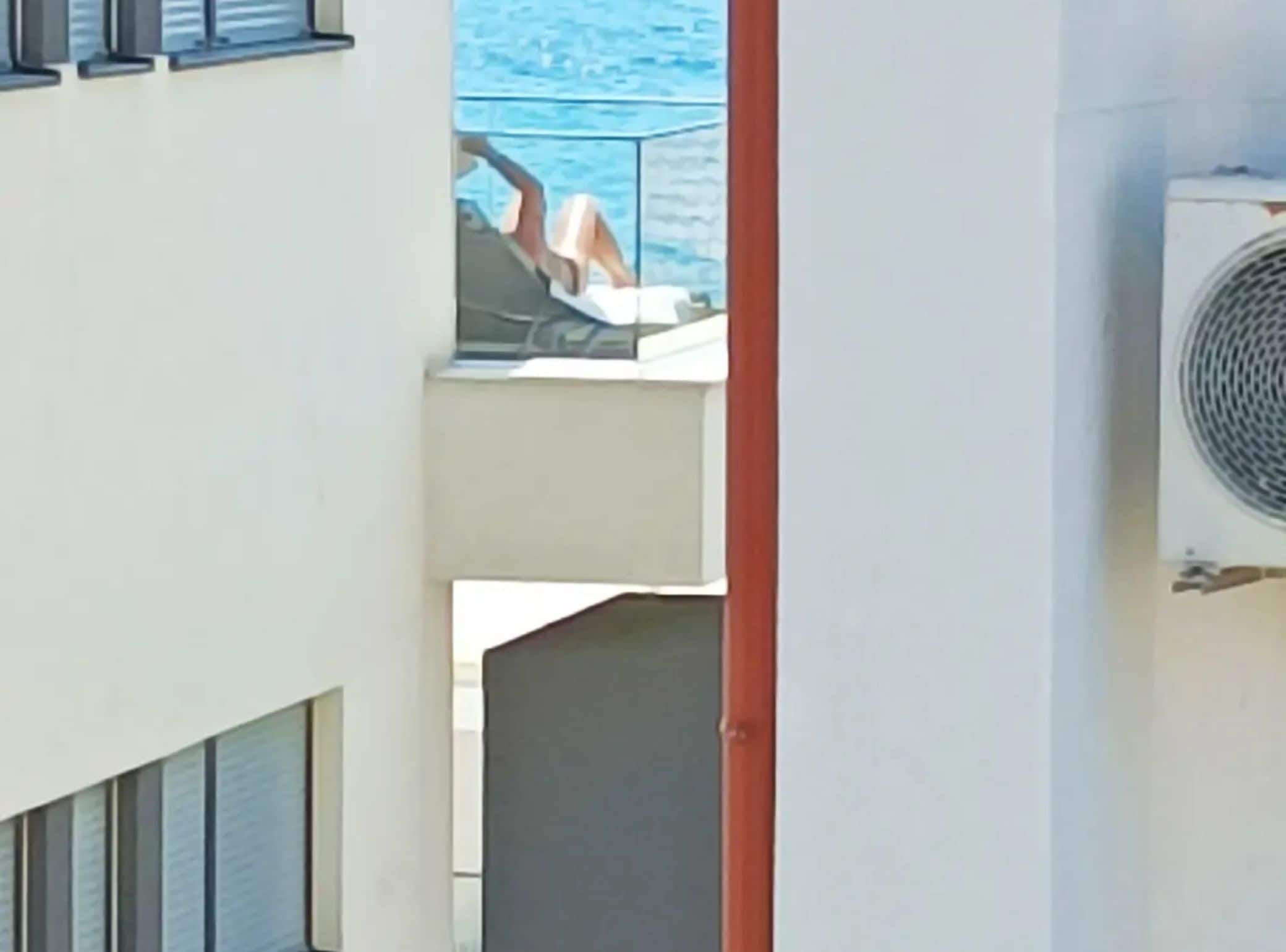 A balcony with a view of the sea, a person relaxing, and an air conditioning unit.