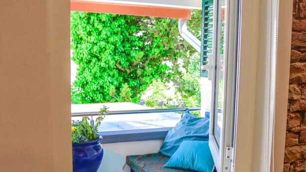 Assento junto à janela com vista para vegetação verde, uma planta em vaso e uma almofada estampada.