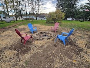 A backyard with a fire pit surrounded by four Adirondack chairs in various colors.