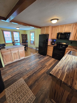A kitchen with wooden cabinets, a black refrigerator, a microwave, and a stove. There is a window with blinds and a rug on the floor.