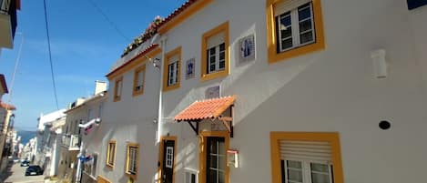 A street view of a building with a yellow and white facade, multiple windows, and a red-tiled roof.