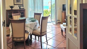 A dining area with a table set for four, chairs, a cabinet with glass doors, a framed picture on the wall, and a view of the sea through a glass door. - Apartment Ora al Mare - 32002 (Pedreña)