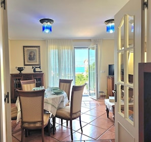 A dining area with a table set for four, chairs, a cabinet with glass doors, a framed picture on the wall, and a view of the sea through a glass door. - Apartment Ora al Mare - 32002 (Pedreña)