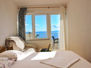 Ein Schlafzimmer mit Bett, Stuhl, Laptop und Meerblick durch ein Fenster.