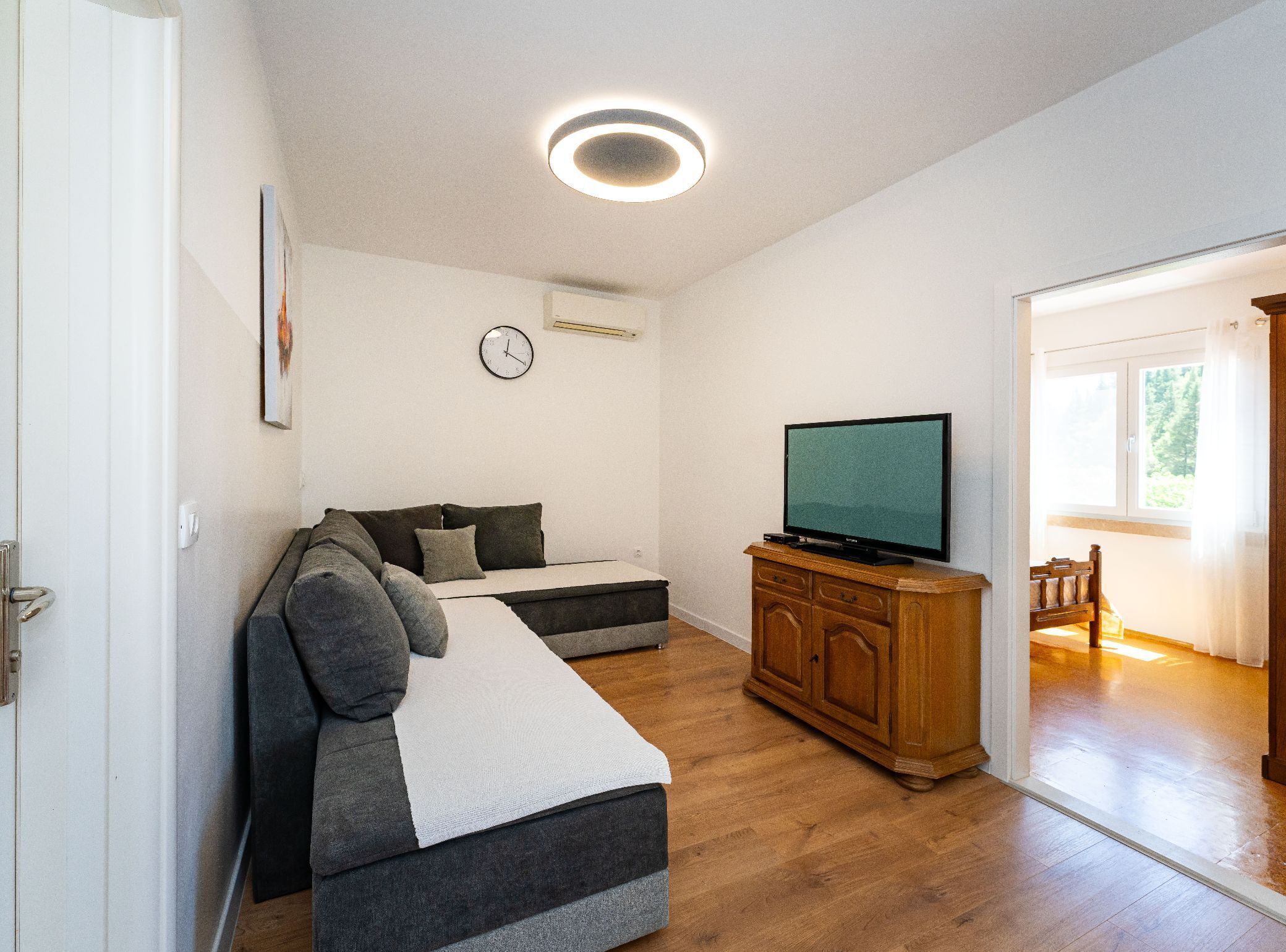 A living room with a sofa, a television on a wooden cabinet, a clock on the wall, and a door leading to another room.