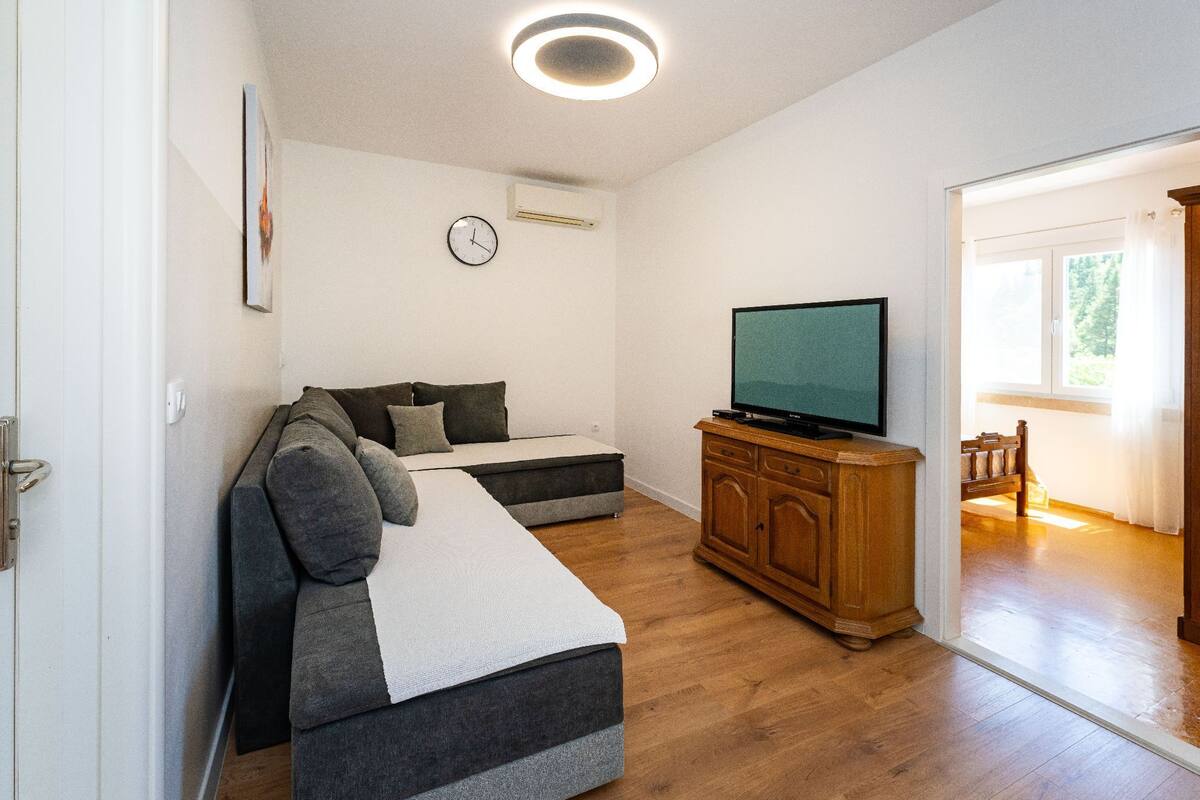 A living room with a sofa, a television on a wooden cabinet, a clock on the wall, and a door leading to another room.