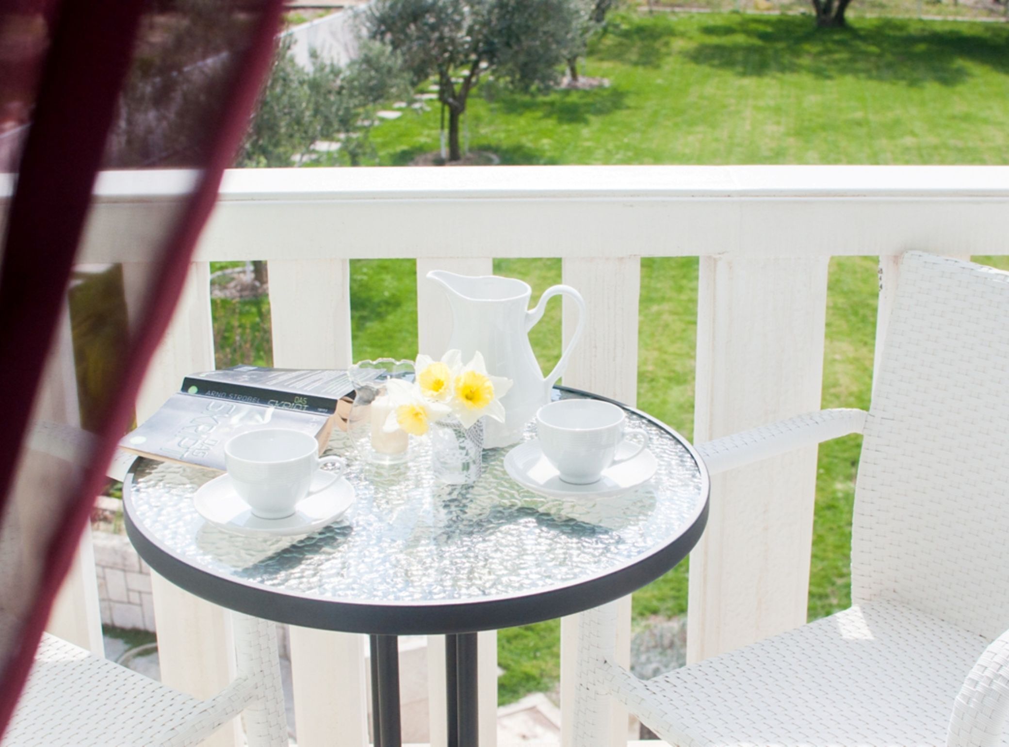 A round glass table with white cups and a pitcher, set on a white deck with a view of a grassy area.