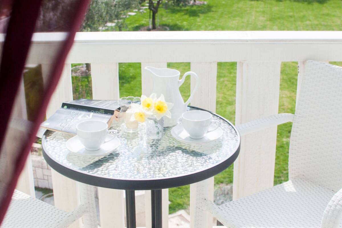 A round glass table with white cups and a pitcher, set on a white deck with a view of a grassy area.