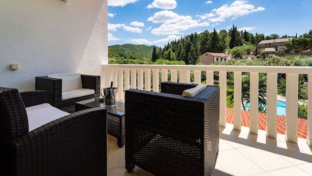 Ein Balkon mit Korbmöbeln, einem Tisch mit zwei Weingläsern und Blick auf eine grüne Landschaft mit Häusern.