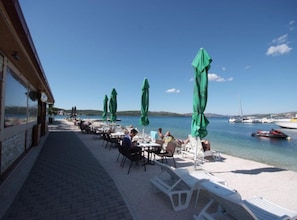 A seaside restaurant with outdoor seating, green umbrellas, and a view of the sea and boats. - Apartments Gogi (Okrug)