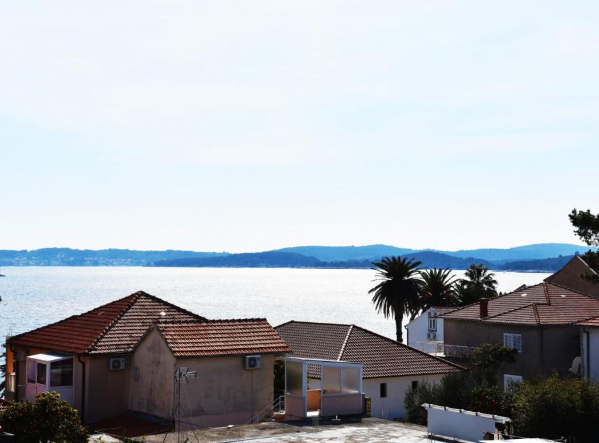 Ein Küstenwohngebiet mit Häusern, einem kleinen Gebäude und Blick auf das Meer und ferne Hügel.