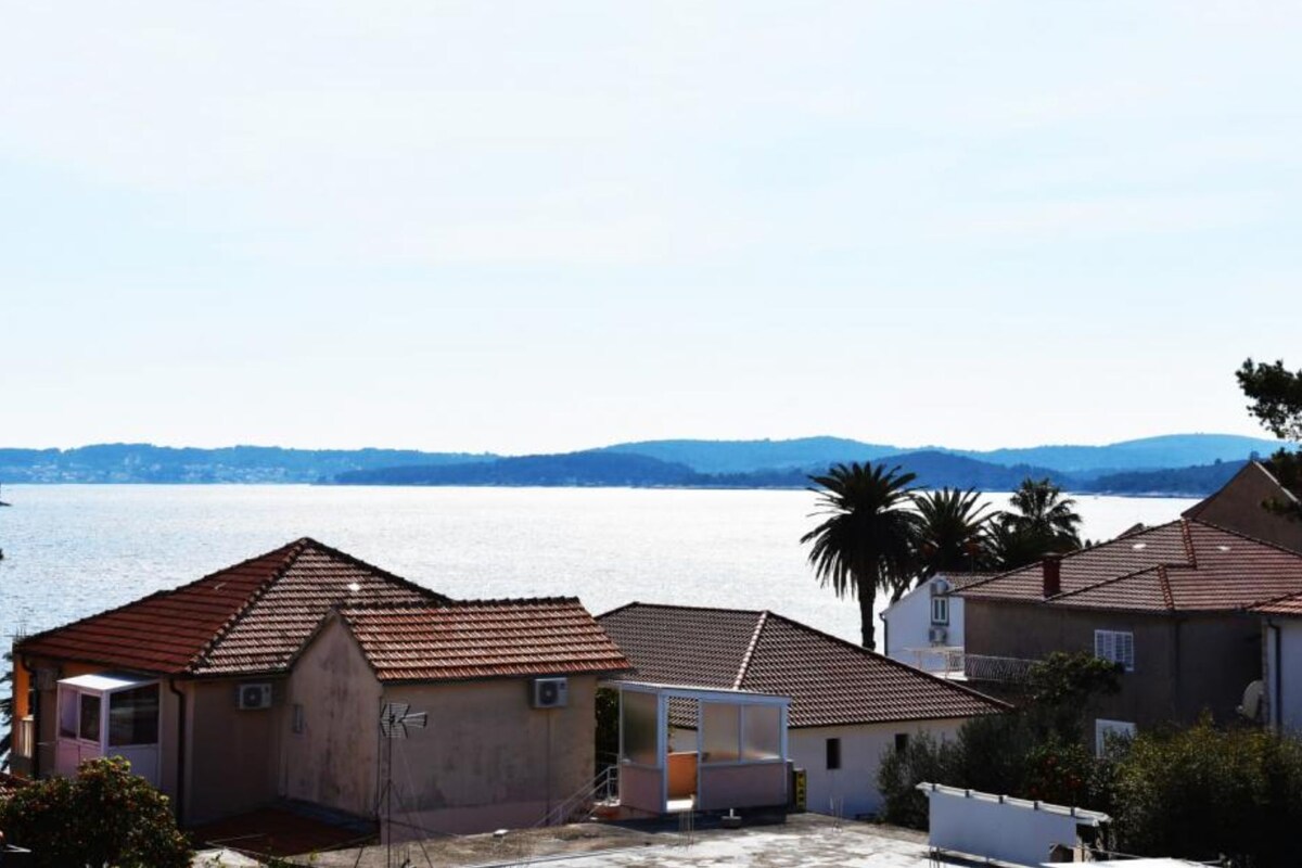 Ein Küstenwohngebiet mit Häusern, einem kleinen Gebäude und Blick auf das Meer und ferne Hügel.