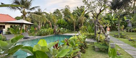 Una piscina rodeada de abundante vegetación, un sendero y un edificio con techo rojo.