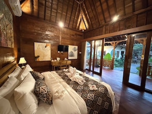 A wooden room with a bed, a desk, and a TV. There are artworks on the walls and a view of the outside. - Laksmi Viridis Villas (Gianyar)