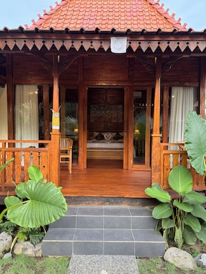 A wooden house with a tiled roof, a porch with a bench, and a door leading to a room with a bed. - Laksmi Viridis Villas (Gianyar)