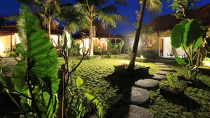 Un jardín tropical con palmeras, un sendero de piedra y edificios tradicionales con techos de tejas.