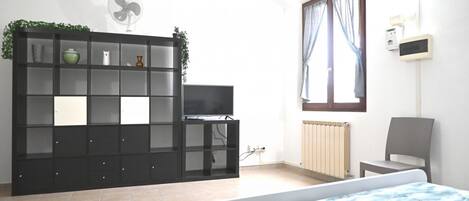 A bedroom with a bed, a TV, a radiator, a fan, and a window with curtains.