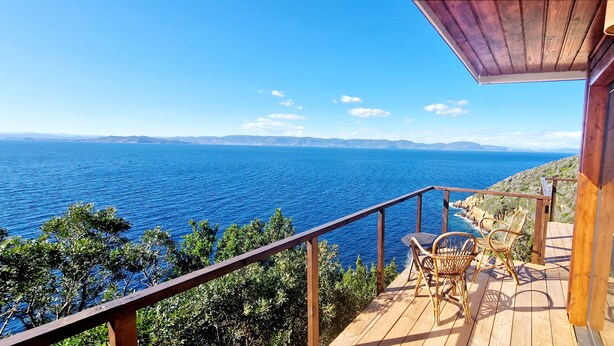 Eine Holzdielen-Terrasse mit Blick auf das Meer, eine Glastür und zwei Korbstühle.