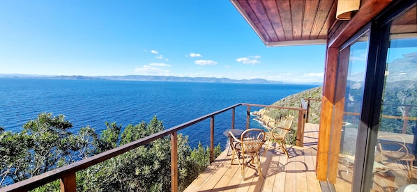 Eine Holzdielen-Terrasse mit Blick auf das Meer, eine Glastür und zwei Korbstühle.