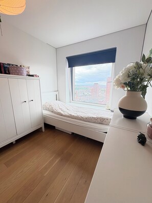 Et soveværelse med seng, et vindue der ud til bygninger, et hvidt klædeskab og en vase med blomster.
