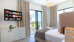 Ein modernes Hotelzimmer mit einem großen Flachbildfernseher, der eine Netflix-Oberfläche zeigt, einem Bett mit einer beige farbenen Decke, einer weißen Kommode und einem Balkon mit Blick.