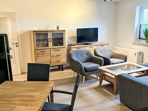 Ein modernes Wohnzimmer mit einem Holzkabinett, zwei grauen Sesseln, einem Holzesstisch und einem Flachbildfernseher.