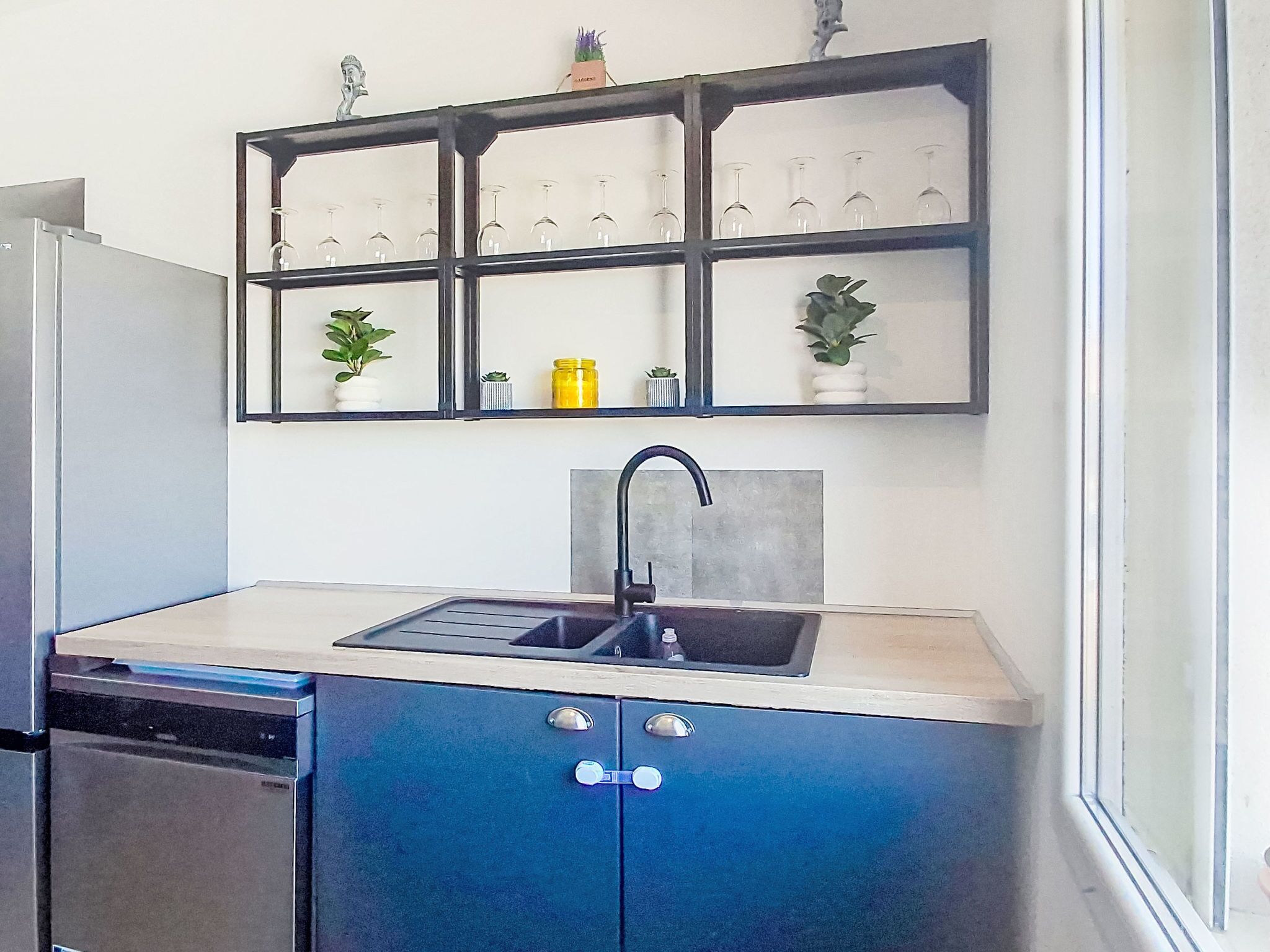 Una cocina con lavavajillas incorporado, un lavamanos y una repisa sobre la encimera con copas de vino y pequeñas macetas.