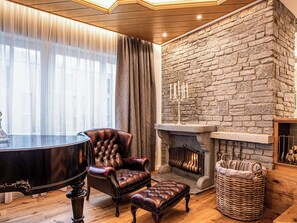 A room with a piano, leather armchair, ottoman, stone fireplace, and a large window with sheer curtains. - Villa 10 by Interhome (Ötz)