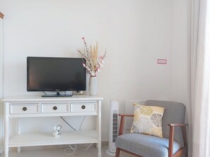 Een witte entertainmentkast met een flatscreen televisie, een vaas met gedroogde bloemen, een radiator en een grijze fauteuil met een gestreept kussen.