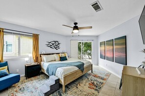 Um quarto moderno com cama, mesas de cabeceira, ventilador de teto, janela grande e obras de arte na parede.