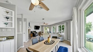 Een ruime woonkamer met een eettafel, stoelen, een plafondventilator en uitzicht door grote ramen.