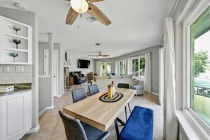 Uma sala espaçosa com mesa de jantar, cadeiras, ventilador de teto e vista através de grandes janelas.
