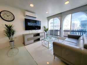 A modern living room with a flat-screen TV, a sofa, a coffee table, and a balcony with a view of buildings. - Da Vinci Suites (San Juan)