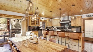 A modern kitchen with wooden cabinets, a large island, bar stools, pendant lights, and a view of the outdoors. - Gold Rush Lodge, 6BR Hot Tub, GameRoom, Sleeps 16+ (Big Bear Lake)