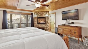 A bedroom with a large bed, a stone fireplace, a wooden ceiling, and a television on the wall. - Gold Rush Lodge, 6BR Hot Tub, GameRoom, Sleeps 16+ (Big Bear Lake)