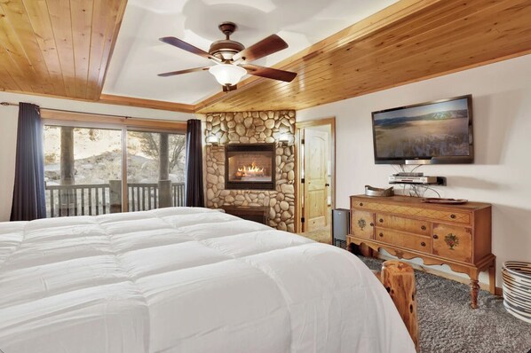 A bedroom with a large bed, a stone fireplace, a wooden ceiling, and a television on the wall. - Gold Rush Lodge, 6BR Hot Tub, GameRoom, Sleeps 16+ (Big Bear Lake)