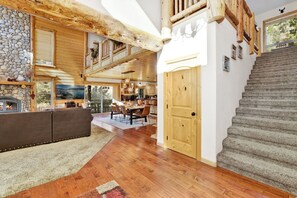 A spacious living area with a fireplace, wooden beams, a dining area, and a staircase. - Gold Rush Lodge, 6BR Hot Tub, GameRoom, Sleeps 16+ (Big Bear Lake)