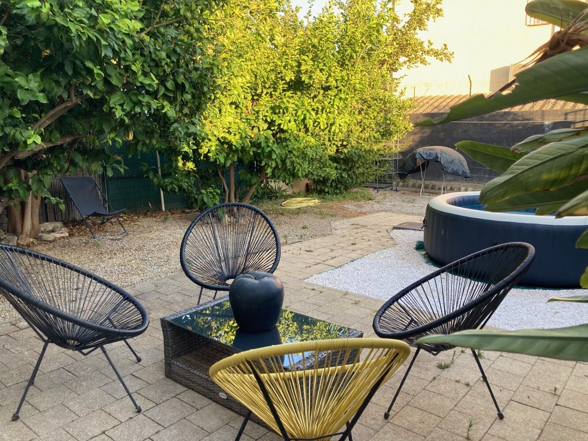 Un espace extérieur aménagé avec des chaises en osier et une table à plateau de verre, entouré de verdure et d’une piscine.