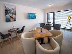A modern room with a wooden table, four chairs, a TV mounted on the wall, and a window with blinds. - V Šachu by Interhome (Dačice)