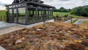 En moderne bygning i glas med et metal tag, omgivet af et naturligt landskab med varieret vegetation.