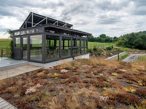 A modern glass structure with a metal roof, surrounded by a natural landscape with diverse vegetation. - V Šachu by Interhome (Dačice)