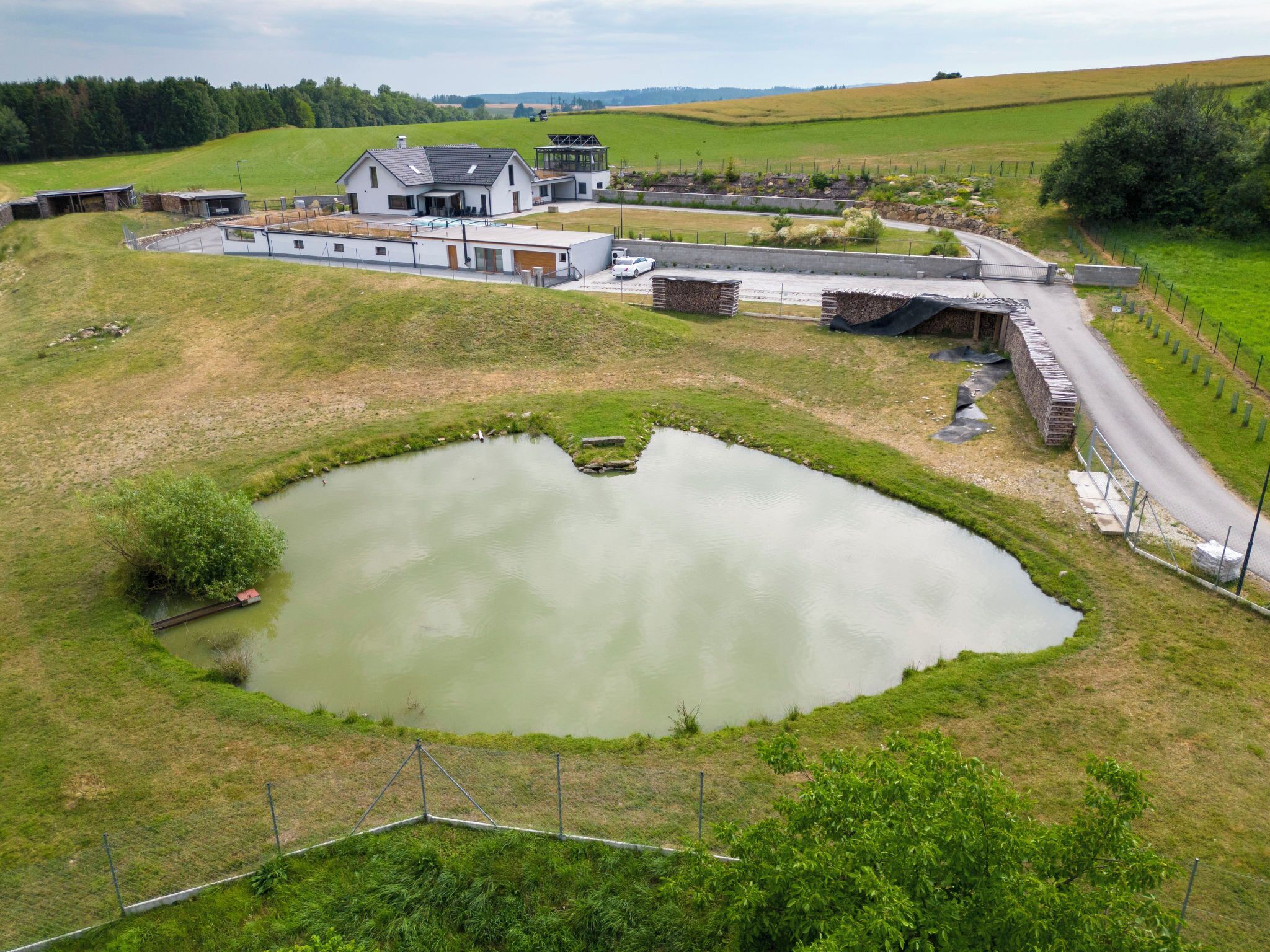 A modern house with a large pond, surrounded by a fenced area and a road.