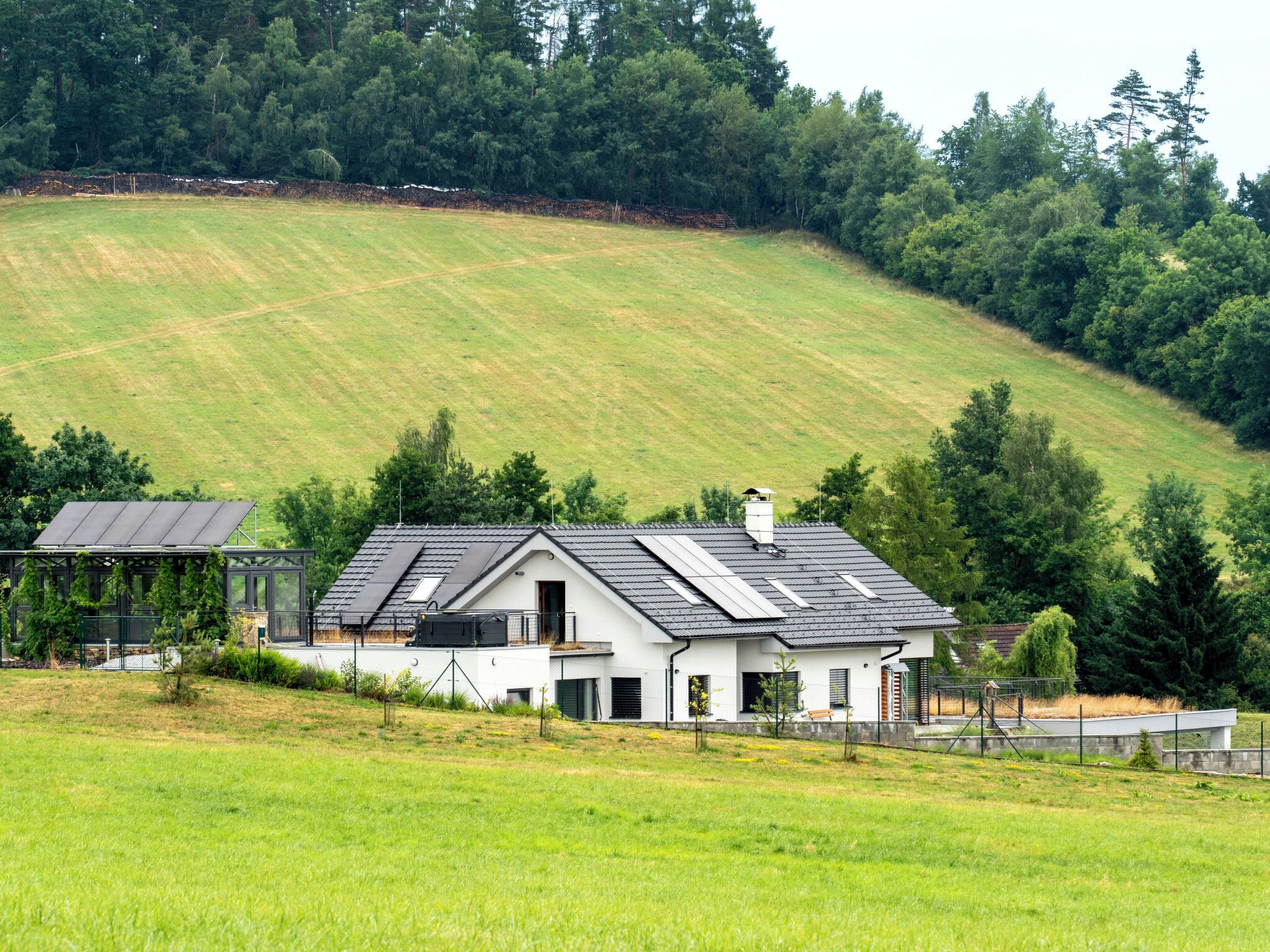 Une maison moderne avec un toit en pente, des panneaux solaires et une vue sur un champ verdoyant et une forêt.