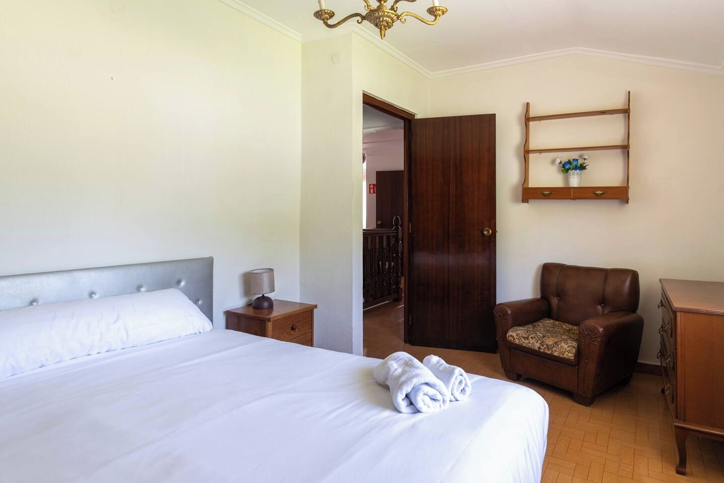 Habitación de hotel con cama, una silla, mesita de noche, armario de madera y estante con un jarrón.