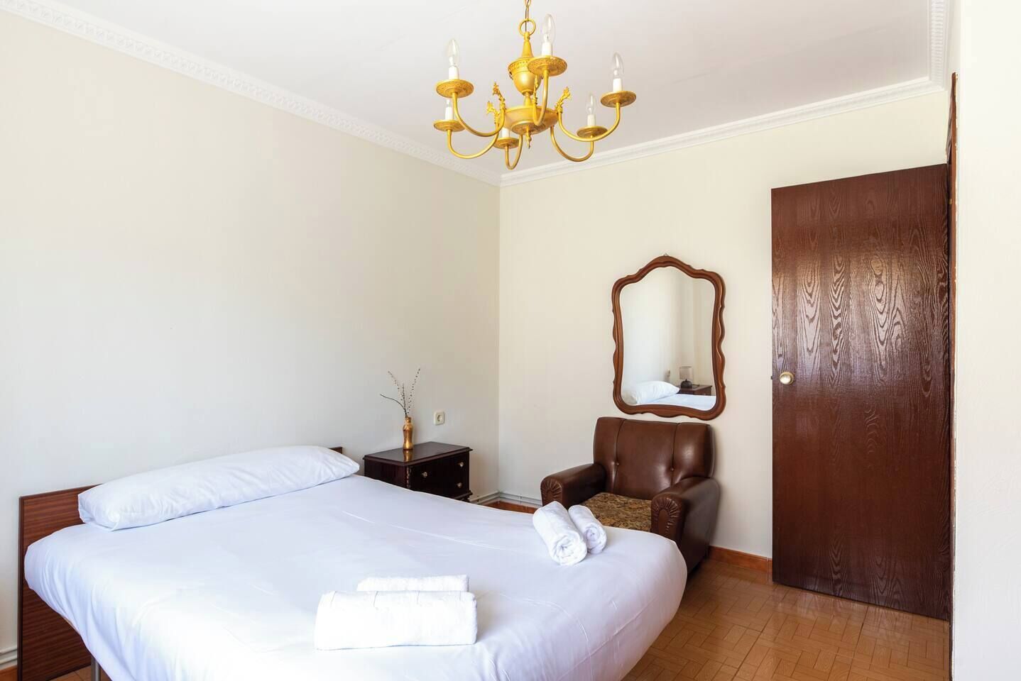 A hotel room with a double bed, a wooden nightstand, a chair, a mirror, and a wooden door.