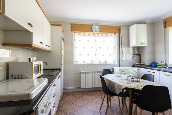 A compact kitchen with a microwave, stove, and sink, a dining table with chairs, and a window with a clock.