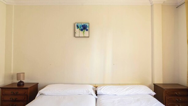 A room with two beds, a nightstand with a lamp, and a painting on the wall. - Casa Pesquera - by Wehomes (Pesquera)